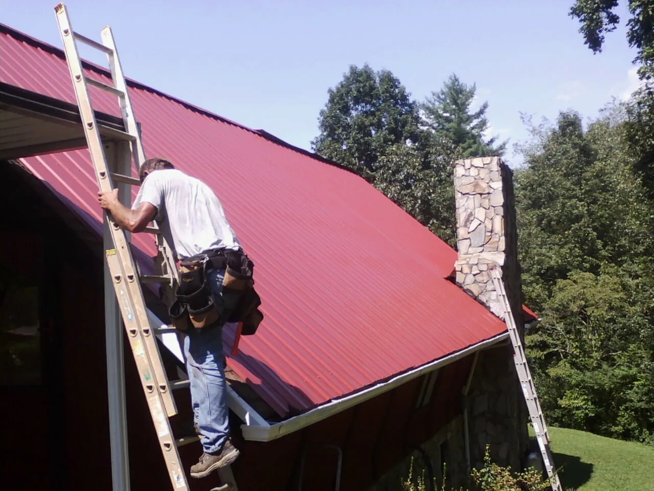 Roofer on a ladder beside a red metal roof and stone chimney — Soffit & Fascia Repair by Roofing Brothers in Hamilton